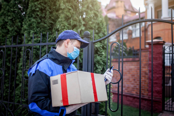A man in service uniform wearing medical mask and gloves delivering a cardboard box to the door of the modern house, outdoor lifestyle, self-isolation in lockdown, contactless online delivery - Photo, Image