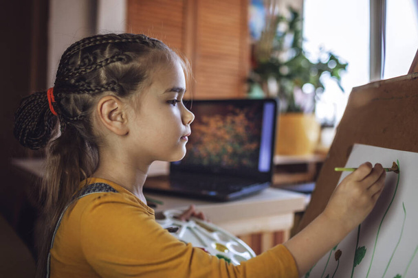 Ein nettes Mädchen mit Palette, Staffelei und Pinsel zeichnet sonnige Blumen, die auf dem Bildschirm des Laptops abgebildet werden, als wäre sie an der frischen Luft, Selbstisolierung und Lebensstil in Quarantäne-Zeit, Online-Workshop - Foto, Bild
