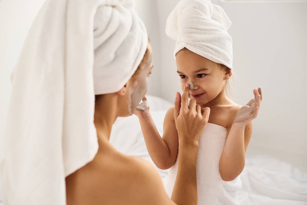A young caucasian mother and little daughter with wrapped hair in white bath towels apply a clay mask on the mother faces - Фото, зображення