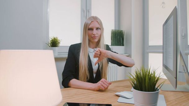 Kobieta z kciukami w biurze. Siedząc w miejscu pracy młoda blondynka w garniturze biznesowym patrząc w kamerę i robiąc kciuki w dół gest dezaprobaty lub niechęci. - Zdjęcie, obraz