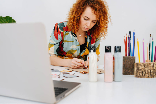 Focused artist woman painting plywood figures behind work desk with a brush - Fotó, kép