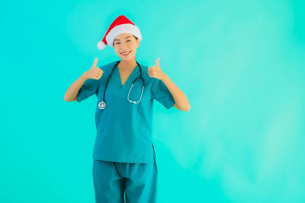 Πορτρέτο όμορφη νεαρή Ασιάτισσα γιατρός γυναίκα φορούν χριστουγεννιάτικο καπέλο με χαμόγελο ευτυχισμένη στο μπλε απομονωμένο φόντο - Φωτογραφία, εικόνα