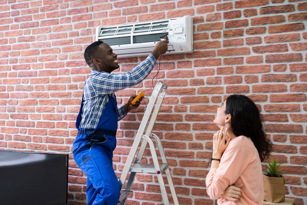 Young Woman Looking At Male Technician Repairing Air Conditioner Mounted On Brick Wall - Foto, Imagem