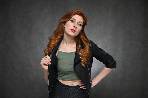 Horizontal portrait of a red-haired young woman in a black jacket and green t-shirt. Model with excellent makeup posing on a gray background in the studio. - Photo, image