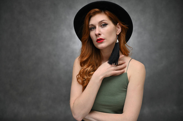 Model in a hat with great makeup posing in a green T-shirt in the studio. Portrait on a gray background of a pretty red-haired young woman. - Fotoğraf, Görsel