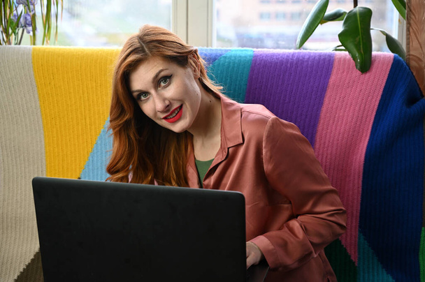 Model is sitting on the sofa with a smile in the home interior. Photo of a red-haired young woman in a pink shirt and black pants, working on a laptop. - Fotografie, Obrázek