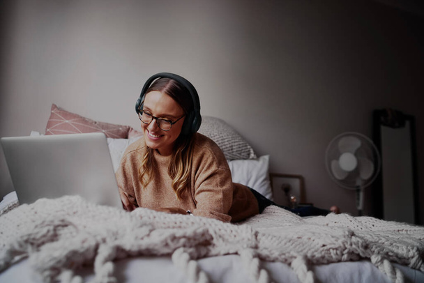 Junge Frau mit Brille trägt Kopfhörer, hört Musik und nimmt an Online-Kursen teil - junge Studentin bei einem Online-Videoanruf  - Foto, Bild