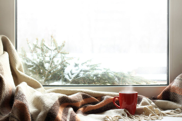 red mug of hot drink on a windowsill with a warm blanket, when behind is window is snow / cozy home atmosphere in the winter - Fotografie, Obrázek