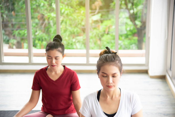 Dos hermosas mujeres asiáticas que están haciendo ejercicio, haciendo yoga en la habitación para practicar sus ejercicios corporales y el control de la respiración son conceptos populares para el mantenimiento del cuerpo
. - Foto, Imagen