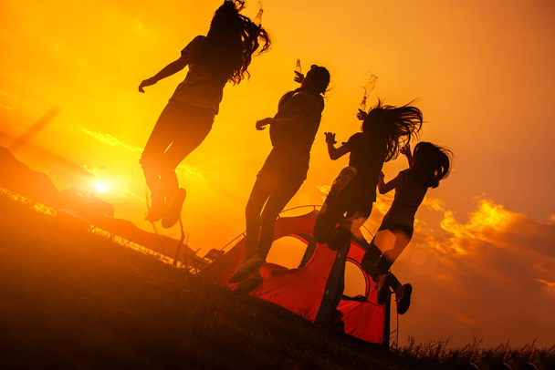 Silhouette Grupo de mujeres de fiesta con botellas de bebidas y saltos disfrutar de viajes de camping, trekking en tiempo de vacaciones al atardecer
. - Foto, Imagen