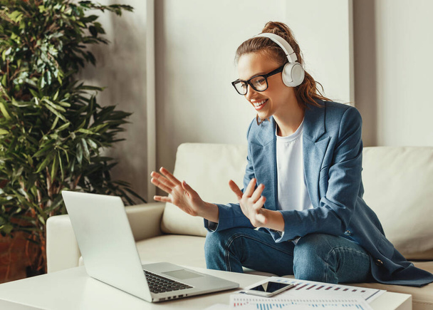Cheerful young female in casual outfit and glasses having video conversation with colleagues and sharing business ideas while sitting on couch in modern workplac - Photo, Image