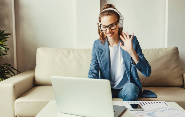 Весела молода бізнес-леді в навушниках проводить онлайн-зустріч з колегами, сидячи на дивані і працюючи з паперами вдома
 - Фото, зображення
