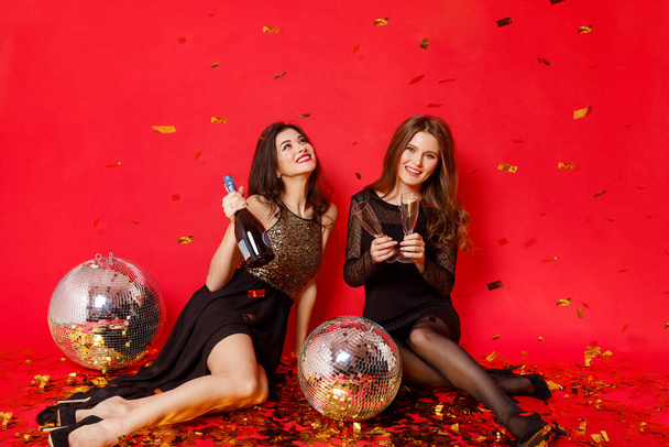 Dos amigas felices en vestidos con maquillaje y labios rojos yacen en el suelo con bolas de disco y confeti de oro sobre un fondo rojo
 - Foto, imagen