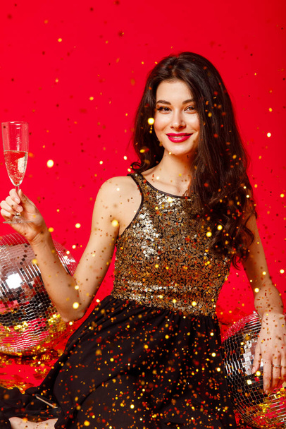 Chica feliz con maquillaje y labios rojos se sienta en el suelo con bolas de discoteca en un vestido negro con lentejuelas de oro y bebidas champán, confeti de oro volar sobre un fondo rojo
 - Foto, imagen