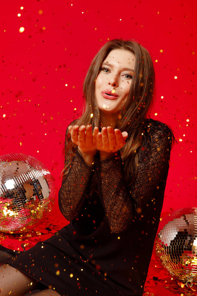 Gelukkig meisje met make-up en rode lippen zit op de vloer met disco ballen in een zwarte jurk en blaast confetti, gouden confetti vliegen op een rode achtergrond - Foto, afbeelding