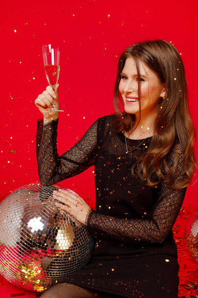Happy girl with make-up and red lips sits on the floor with disco balls in a black dress and drinks champagne, gold confetti fly on a red background - Fotografie, Obrázek