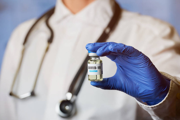 Un médecin spécialiste des maladies infectieuses porte un manteau blanc et un stéthoscope tenant à la main dans un gant médical jetable un vaccin contre le coronavirus (COVID-19). Un médecin avec un vaccin dans un hôpital
. - Photo, image