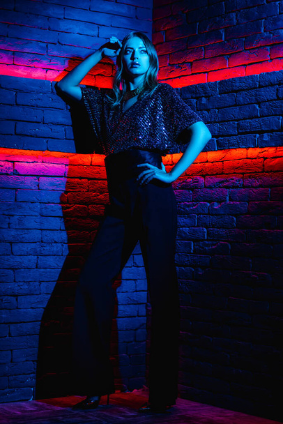 Mooi meisje model in neon verlichting. Jonge vrouw in een jurk met een open rug in rood met blauwe verlichting in een club - Foto, afbeelding