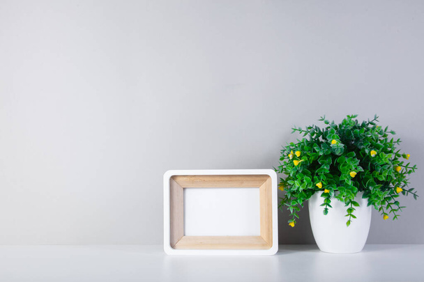 White photo frame on a white table and a wall. A vase of green flowers next to the frame. Copy space of a white wall. - Photo, image