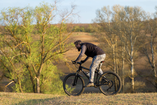 Sports brutal bearded guy on a modern mountain bike. Cyclist on the green hills in the spring. - Foto, imagen