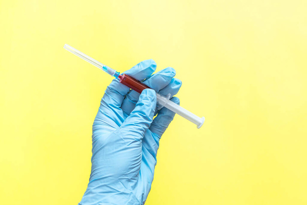 Human hand in rubber glove and disposable syringe with blood samples on yellow background. Concept of coronavirus, COVID-19 pandemic, global quarantine. - Photo, Image