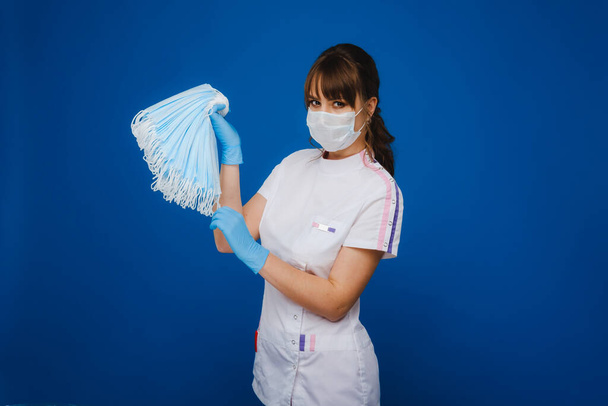 A young girl doctor in a medical mask holds a lot of masks in her hands. A nurse poses on a blue background. - Фото, изображение