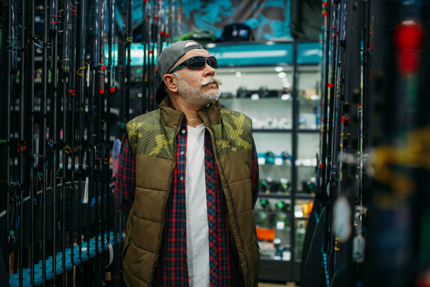 Fisherman in sunglasses choosing rod in fishing shop. Equipment and tools for fish catching and hunting, accessory choice on showcase in store - Fotografie, Obrázek