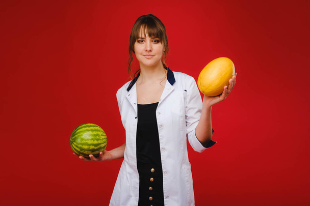 a female doctor nurse in a white coat with fruit in her hands poses on a red background, melon, watermelon - Foto, Imagem