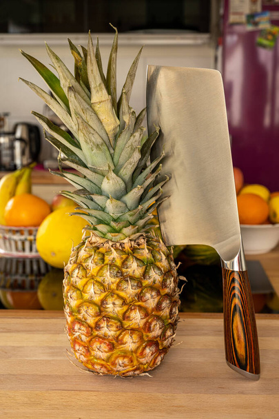 Close-up view of a whole fresh whole pineapple fruit with a cleaver on a cutting board in a domestic kitchen with other varieties of fruit in the background - Foto, Imagen