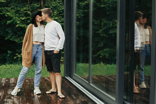 Stylish hipster couple hugging and kissing on wooden terrace, relaxing in cabin in mountains. Happy young family in modern outfits embracing on background of big window and trees - Photo, Image