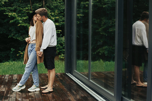 Stylowa para hipsterów przytula się i całuje na drewnianym tarasie, relaksując się w chatce w górach. Szczęśliwa młoda rodzina w nowoczesnych strojach obejmująca na tle dużych okien i drzew - Zdjęcie, obraz