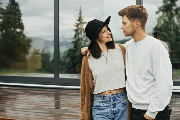 Stylish hipster couple hugging on wooden porch, relaxing in modern cabin in mountains. Happy young family in modern outfits embracing on background of terrace and big window - Photo, Image