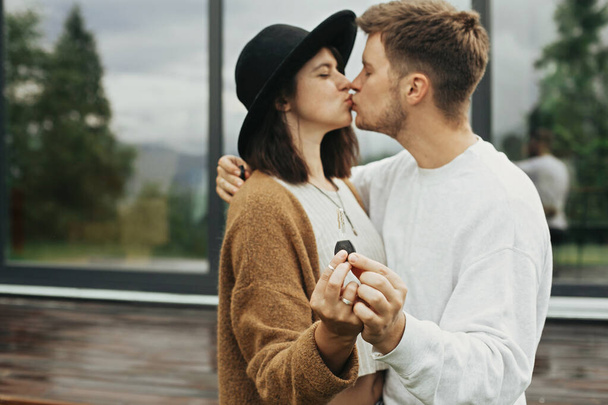 Stylish hipster couple holding together key from their new home on background of terrace and big windows outdoors. Happy young family kissing, celebrating buying first property. - Foto, afbeelding