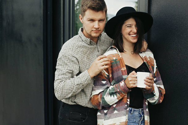 Stylish hipster couple with morning coffee smiling and relaxing on background of modern cabin with big windows in mountains. Happy young family enjoying new home in woods. Space for text - Фото, зображення