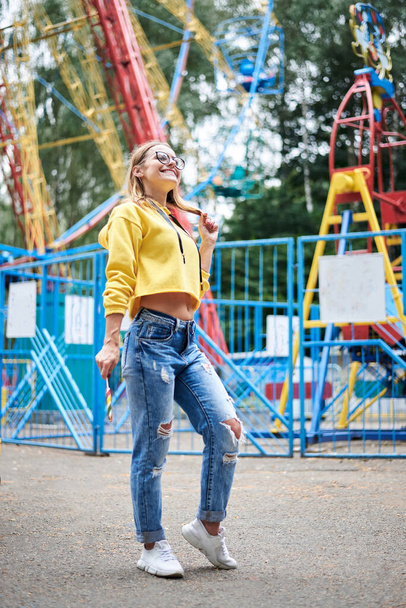 Young blond woman, wearing yellow hoody and blue jeans, holding colorful sweet tasty lollypop in amusement theme park. Full-length portrait of pretty girl, standing in front of ferris wheel. Leisure - Fotó, kép