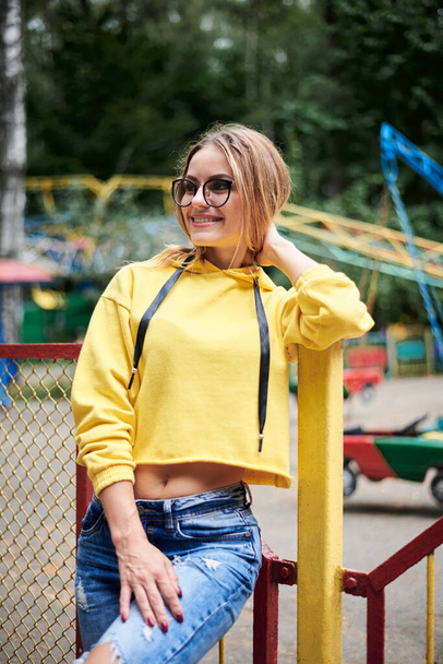 Young blond woman, wearing yellow hoody, blue jeans, spending time in amusement theme park in summer. Three-quarter portrait of pretty girl, leaning on fence railing in front of colorful ferris wheel - Valokuva, kuva
