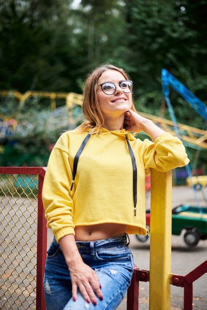 Young blond woman, wearing yellow hoody, blue jeans, spending time in amusement theme park in summer. Three-quarter portrait of pretty girl, leaning on fence railing in front of colorful ferris wheel - Photo, Image