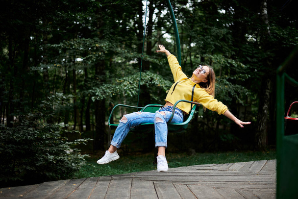 Young blond woman, wearing yellow hoody and blue jeans, riding on carrousel, smiling, laughing in park with green trees in summer. Entertainment in amusement park. Sunday leisure time. - Fotó, kép