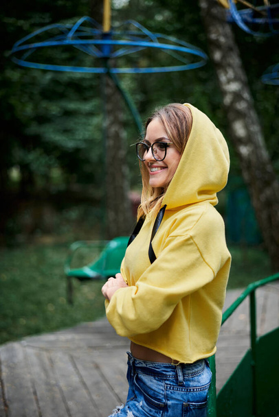 Young blond woman, wearing yellow hoody, blue jeans and eyeglasses, standing on old carrousel in park in summer Three-quarter portrait of girl spending free time in city forest. Summer entertainment. - Фото, зображення