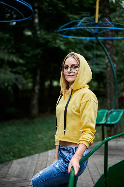 Young blond woman, wearing yellow hoody, blue jeans and eyeglasses, standing on old carrousel in park in summer Three-quarter portrait of girl spending free time in city forest. Summer entertainment. - Foto, Bild