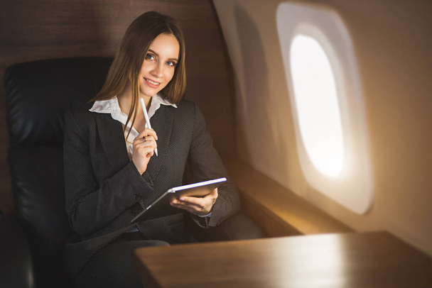 Happy brown haired young caucasian businesswoman holding tablet, sitting on seat in private jet, looking at airplane window and smiling. - Fotó, kép