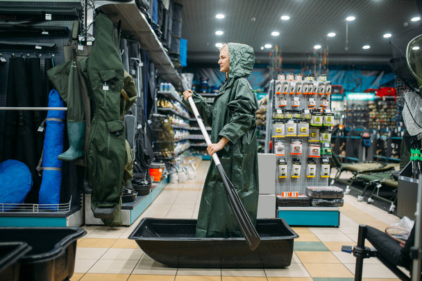 Fisherwoman in a raincoat is sailing on a boat in fishing shop, side view, hooks and baubles on background. Fisherwoman shoosing equipment and tools for fish catching and hunting, accessory in store - Zdjęcie, obraz