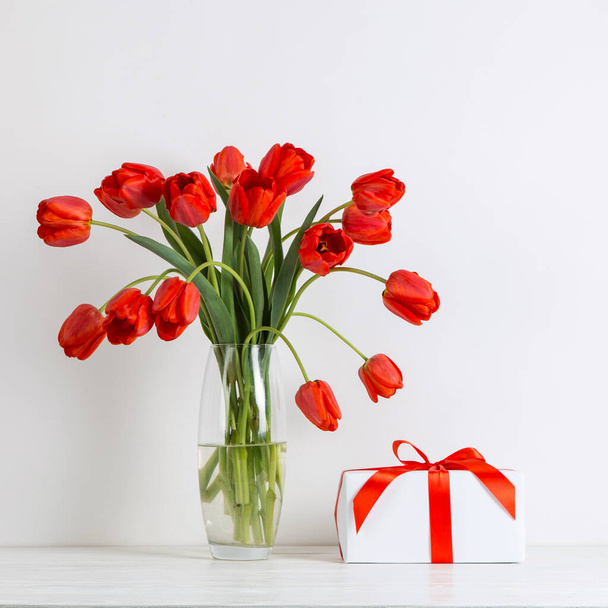 Red tulips in a vase and gift on the table, on a white background. Postcard blank. - Foto, Imagem
