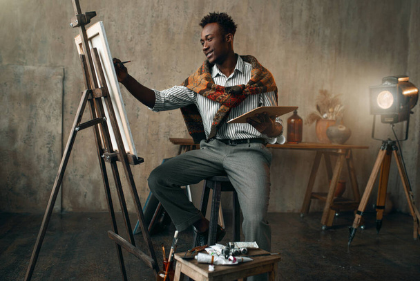 筆とパレットで笑顔の画家は、イーゼルに座っている。男性アーティストは彼の職場で、ワークショップで創造的なマスター作品を描く - 写真・画像
