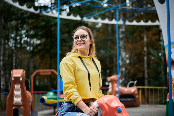 Young blond woman, wearing yellow hoody, blue jeans and eyeglasses, riding carrousel with colorful animals in park with green yellow trees. Adult girl having fun in amusement park, laughing, smiling. - Фото, изображение