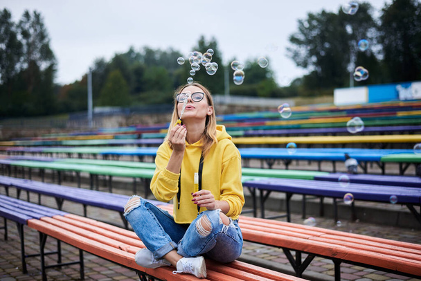 Young blond woman, wearing yellow hoody, blue jeans and eyeglasses, sitting on colorful bench in city urban park in summer. Portrait of pretty girl, blowing making soap bubbles, laughing, smiling. - Fotó, kép