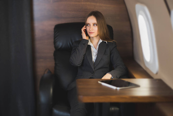 Hermosa mujer rica en avión de primera clase. Joven mujer de negocios en ropa formal sentada en jet privado, hablando por teléfono celular durante el vuelo
. - Foto, Imagen