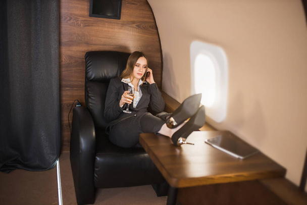 Confident self assured young businesswoman sitting in private plane with her feet on table, talking on mobile phone, holding glass of champagne, wearing formal black suit, high heel shoes. - Fotoğraf, Görsel