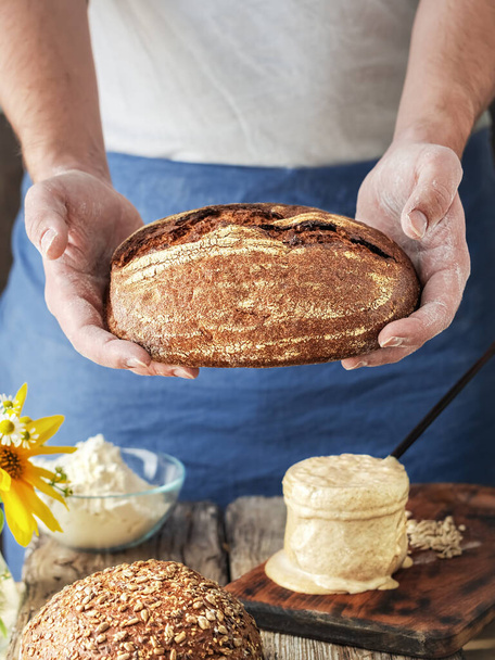 Świeżo upieczony chleb organiczny. Piekarz trzyma w rękach chleb rękodzielniczy domowej roboty, zrobiony z mąki, drożdży, soli i wody. Domowej roboty wypieki w okresie samoizolacji, zakwasie, zbliżenie - Zdjęcie, obraz