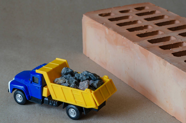 Een gele vuilniswagen draagt puin naast een baksteen. Speelgoedtruck met een volledige opbouw van bouwmaterialen. Creatieve industriële achtergrond. Bouw en vervoer van bouwmaterialen. Selectieve focus. - Foto, afbeelding